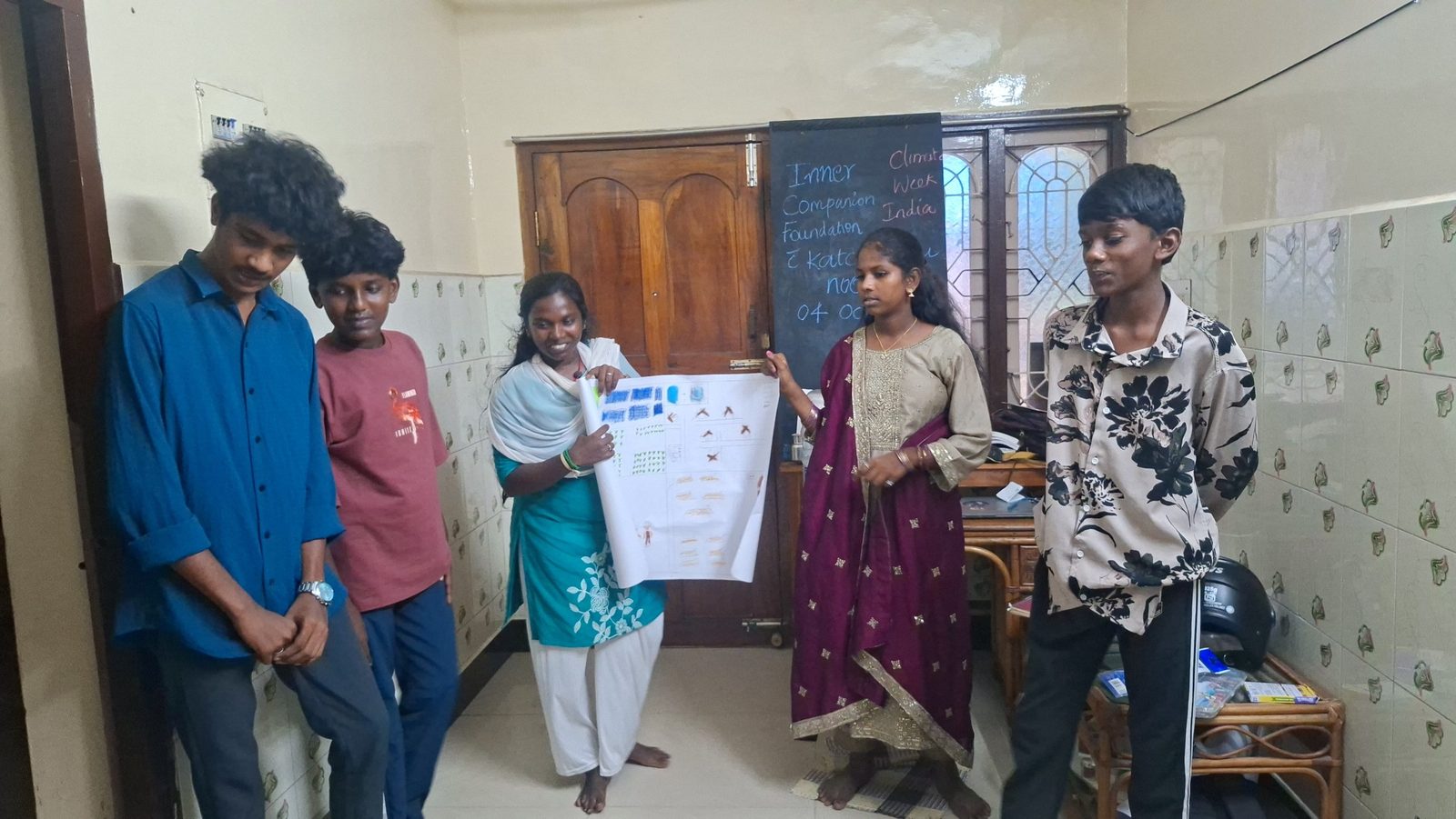 Five young people stand in a room as two of them hold up their drawing of rows of blue panels, plants and birds, beside a chalkboard reading Inner Companion Foundation and Katchipedu Nook, Climate Week India