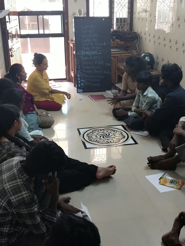 Participants sit in a circle on the floor around a patterned floor medallion. A chalkboard behind them lists words from the conversation: why, reflect, surprise, shock, greed, sad, anger, problem, questions, helplessness, solutions, animals