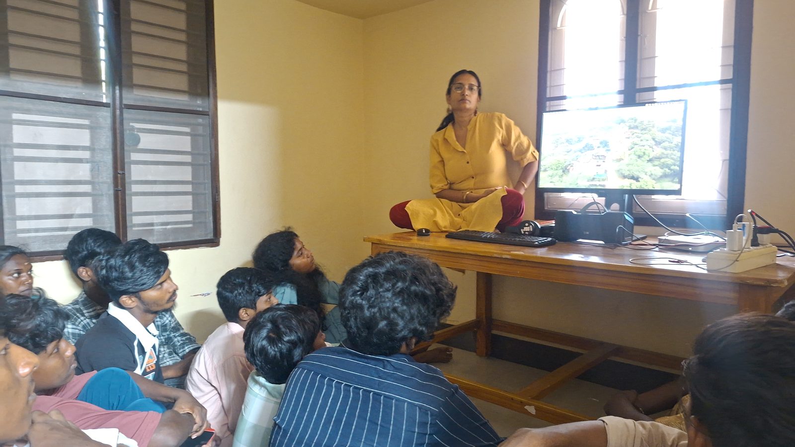 A woman in a yellow kurta sits cross-legged on a table beside a monitor showing a documentary, while young people seated on the floor watch