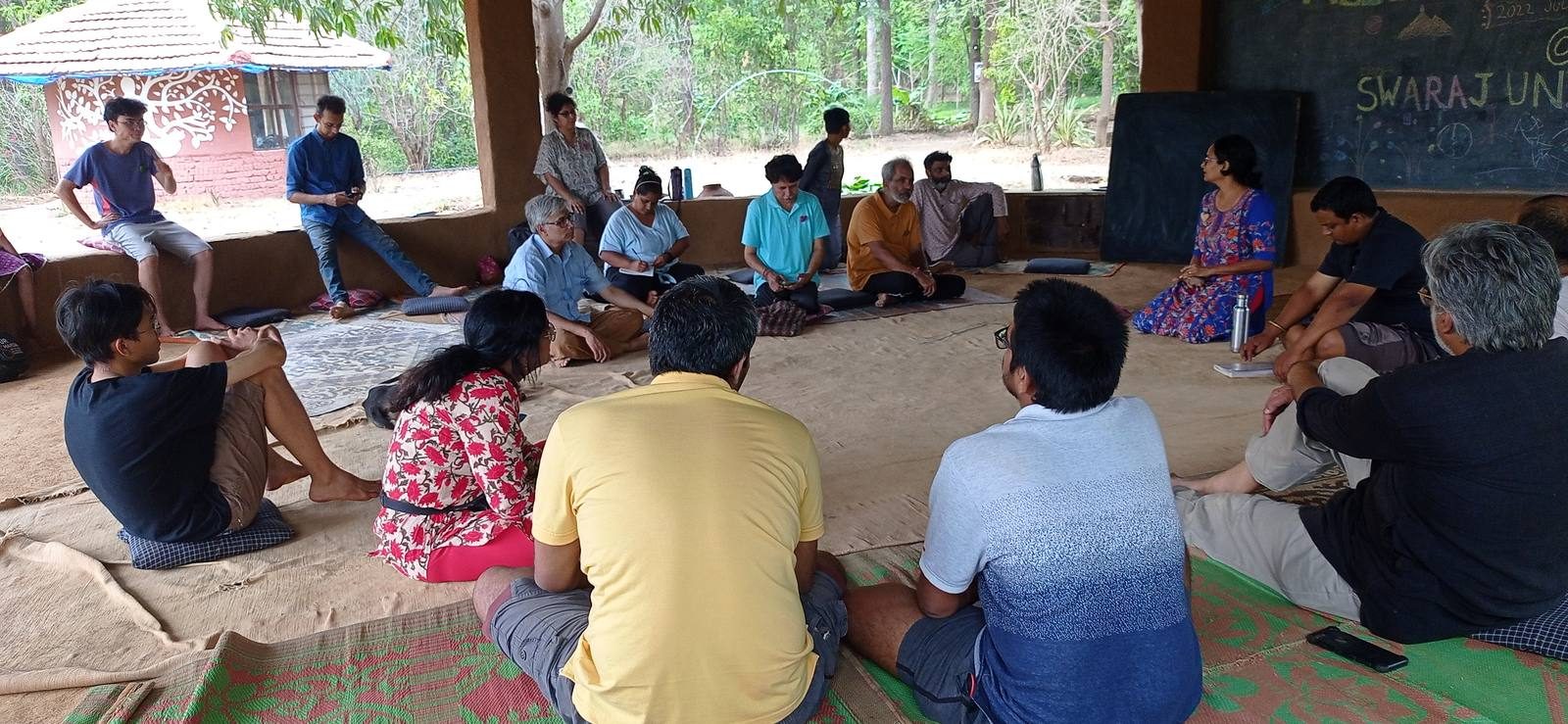 Adults sitting on mats in a circle on the floor of an open-walled hall, working together in discussion.
